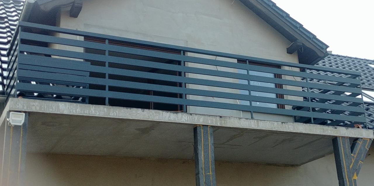 Szara, pozioma balustrada balkonowa z prostokątnych profili, zamontowana na surowym, betonowym balkonie domu jednorodzinnego w trakcie budowy.