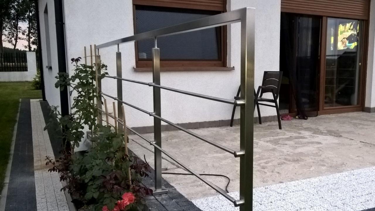 Zewnętrzna balustrada ze stali nierdzewnej z trzema poprzeczkami, zamontowana na tarasie przed domem z białym tynkiem i brązowymi oknami, z roślinami w donicy po lewej stronie.