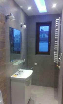 Nowoczesna łazienka z szarymi płytkami, podświetlanym sufitem, umywalką na szafce i grzejnikiem drabinkowym, widok na dom przez okno.