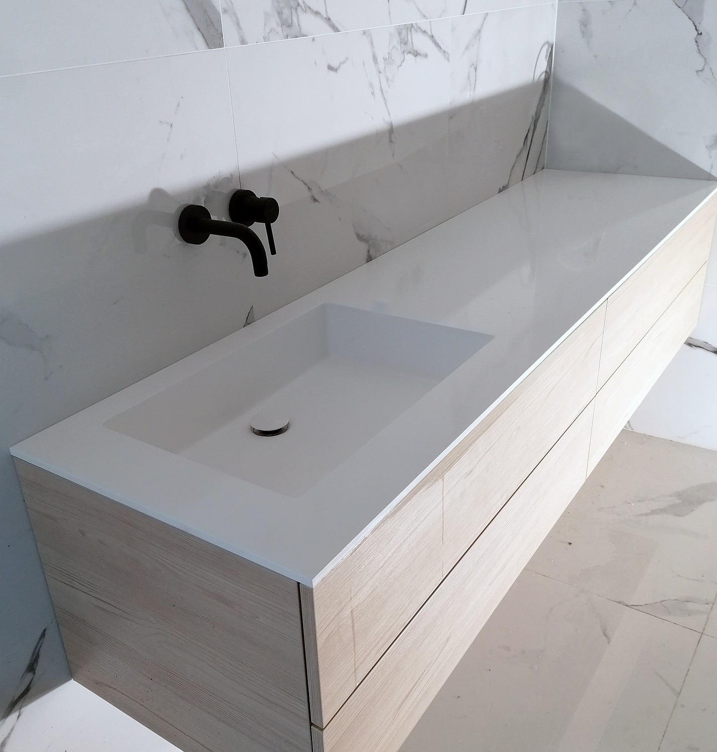 Biały blat łazienkowy ze zintegrowaną umywalką, wykonany z konglomeratu mineralnego solid surface, zamontowany na szafce z frontami imitującymi jasne drewno. Na ścianie widoczna czarna bateria...