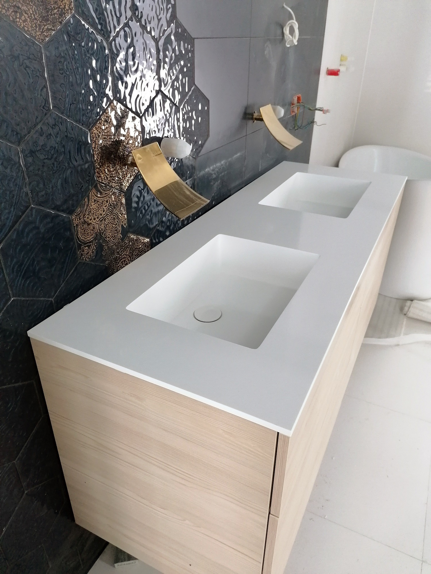 Łazienkowy blat z dwiema prostokątnymi umywalkami wpuszczanymi, wykonany z białego konglomeratu na szafce z jasnego drewna, widoczne złote baterie i heksagonalne płytki na ścianie.