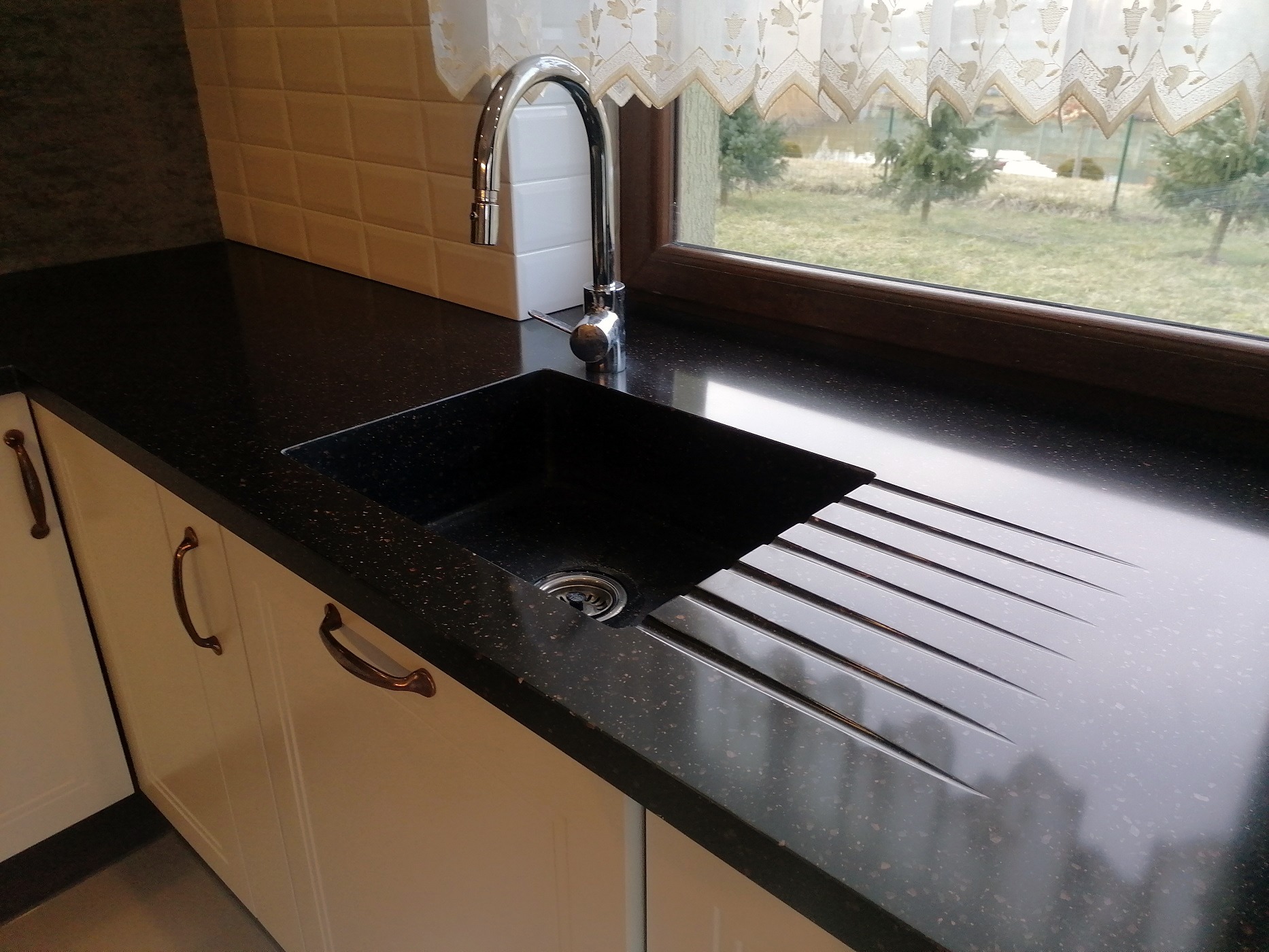 Czarny, błyszczący blat kuchenny z kamienia z wbudowanym zlewem i ociekaczem, widok na okno z firanką i zielenią na zewnątrz.