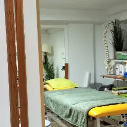 Gabinet z łóżkiem do masażu z zielonym pokrowcem i żółtymi elementami, widok przez drewniane drzwi przesuwne, na ścianie model kręgosłupa i półka z akcesoriami.