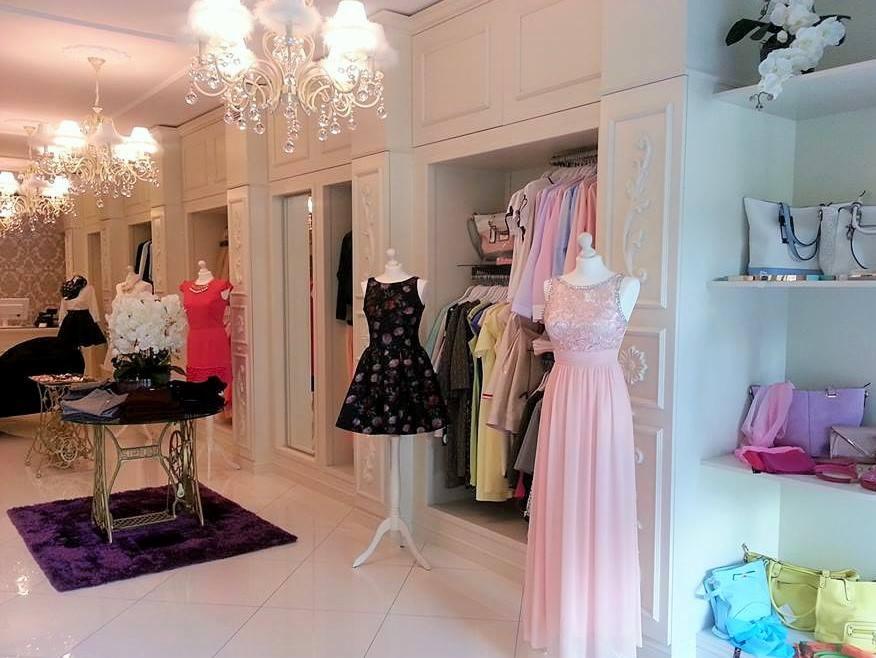 Elegancki butik z sukniami na manekinach, kryształowymi żyrandolami i zdobionymi szafami wnękowymi, z maszyną do szycia przerobioną na stolik kawowy.