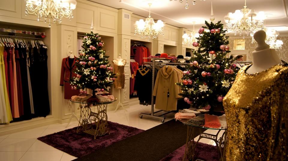 Wnętrze butiku odzieżowego z dekoracjami świątecznymi, manekin w złotej sukience na pierwszym planie, choinki na stolikach ze starych maszyn do szycia.
