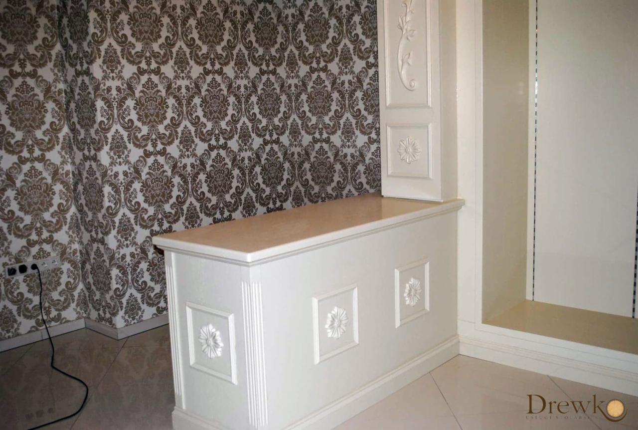 Biała, drewniana szafka z blatem i dekoracyjnymi, kwiatowymi panelami na tle ściany w brązowe ornamenty i fragmentu białej szafy wnękowej.