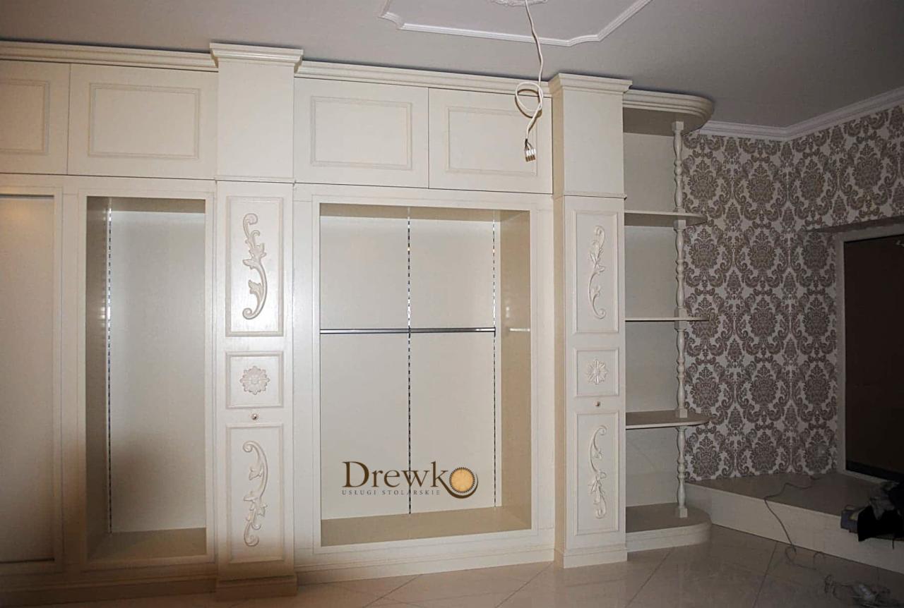 Biała, zabudowana szafa z ornamentami w klasycznym stylu, widoczne półki i drążek na ubrania, na jednej z półek logo firmy stolarskiej.