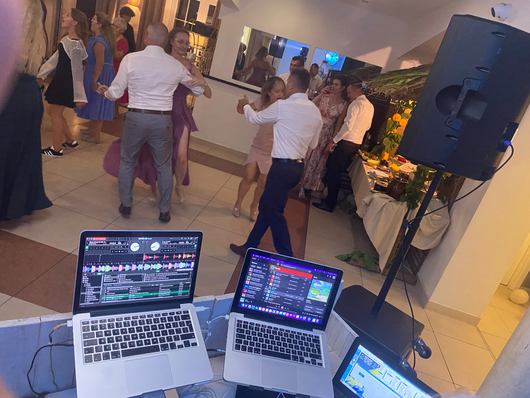 Parkiet taneczny podczas imprezy z DJ-em, widoczne laptopy z oprogramowaniem dla DJ-ów i tańczący goście w tle. Sprzęt nagłośnieniowy ustawiony z boku.