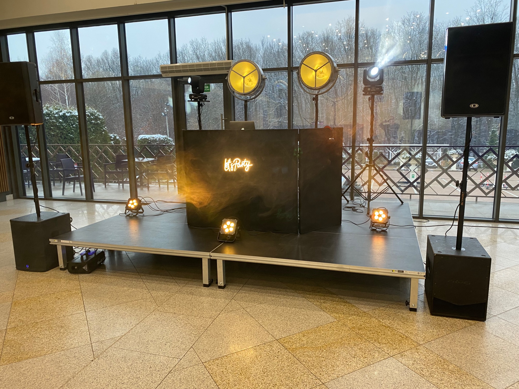 Scena z nagłośnieniem i oświetleniem 'Let's Party' przed oknem z widokiem na zimowy krajobraz. Dwa reflektory punktowe i kolumny głośnikowe po bokach.