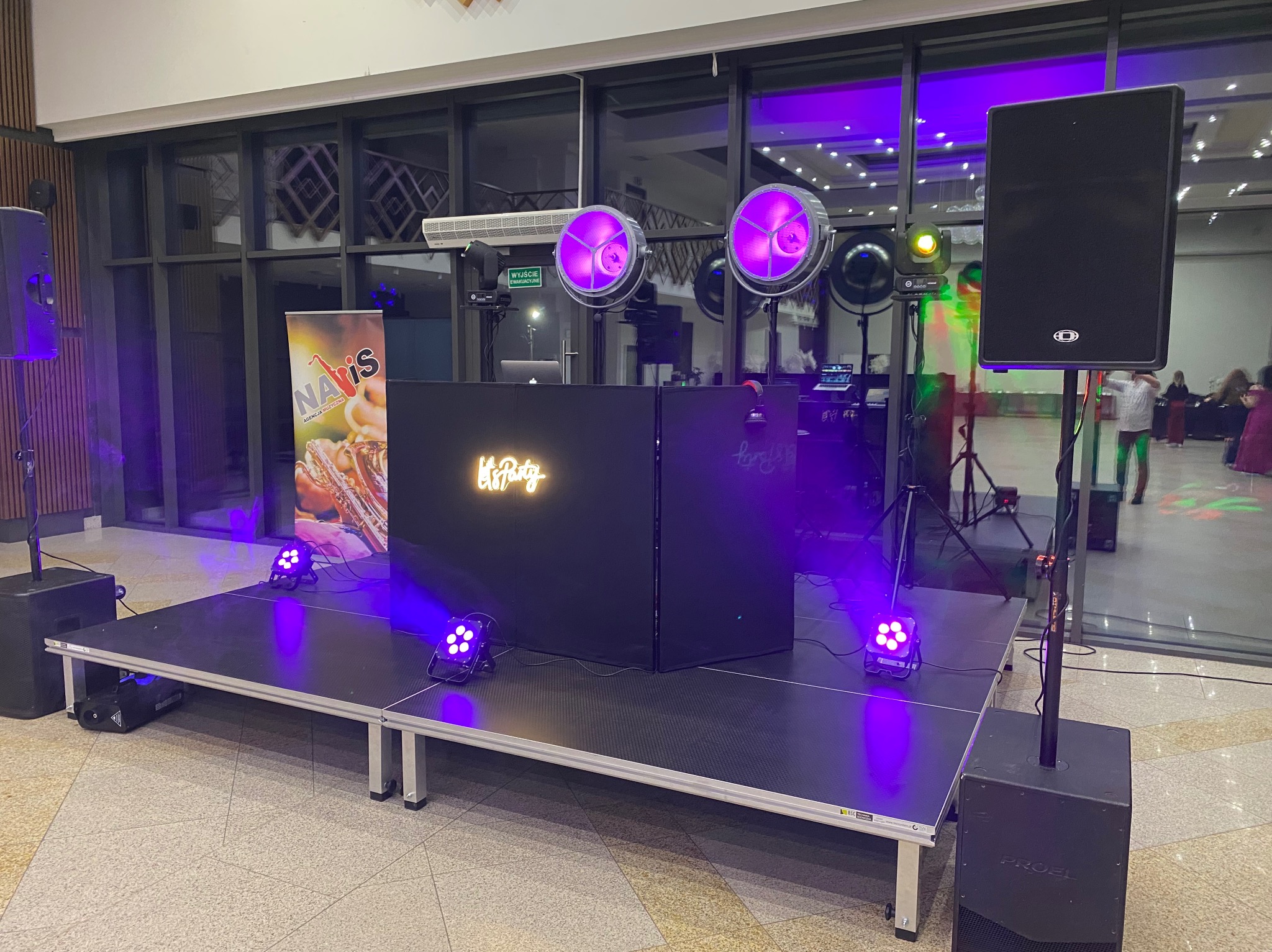 Scena z nagłośnieniem i oświetleniem w fioletowych barwach, przygotowana na występ. Widoczny baner reklamowy i sprzęt DJ-ski. Parkiet taneczny w tle.