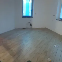 Wnętrze w trakcie remontu: ułożona podłoga z płytek imitujących drewno, okna zabezpieczone folią malarską, worki z zaprawą w tle, widoczny fragment drabiny.