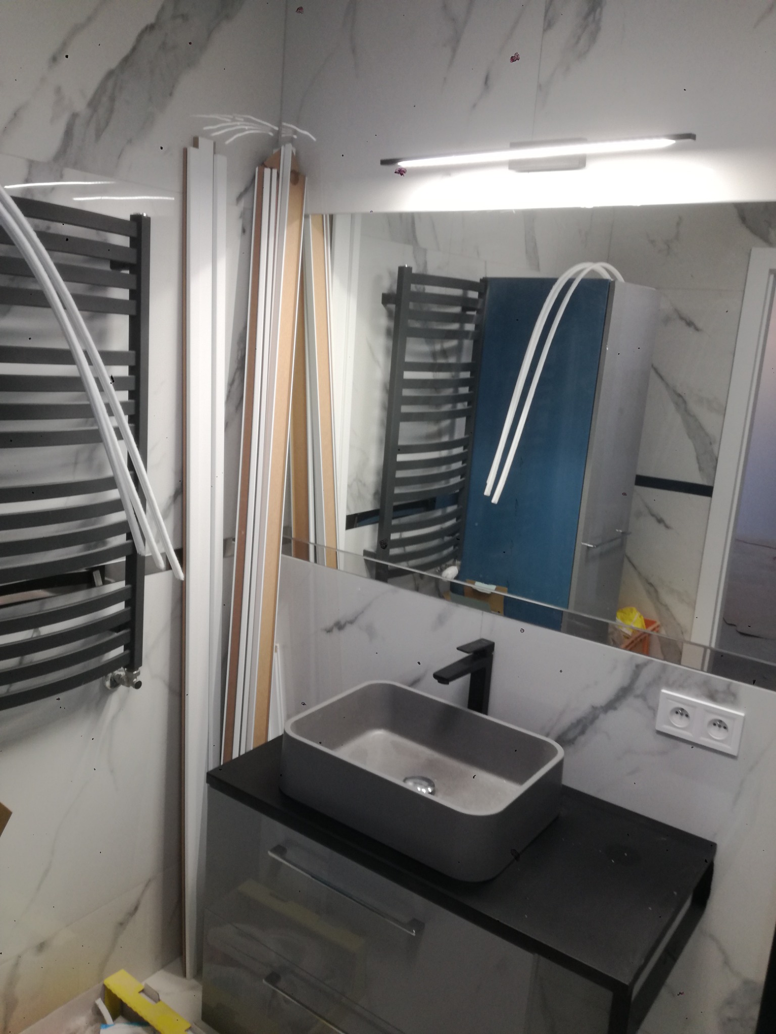 Szara umywalka nablatowa na czarnej szafce w łazience z marmurowymi płytkami, widoczny grzejnik drabinkowy i listwy przypodłogowe przed montażem.