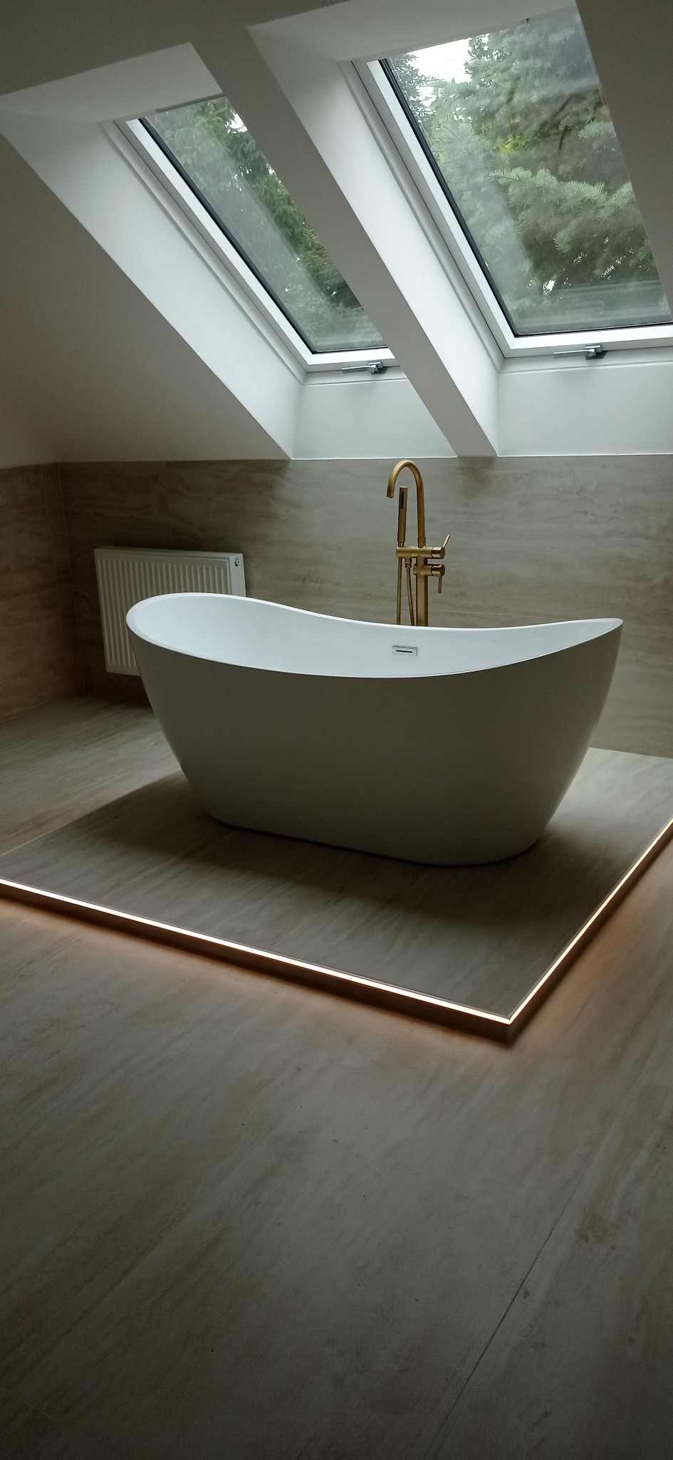 Nowoczesna, wolnostojąca wanna z złotą armaturą, oświetleniem LED pod platformą i oknami dachowymi, w wykończonej łazience w beżowych tonacjach.
