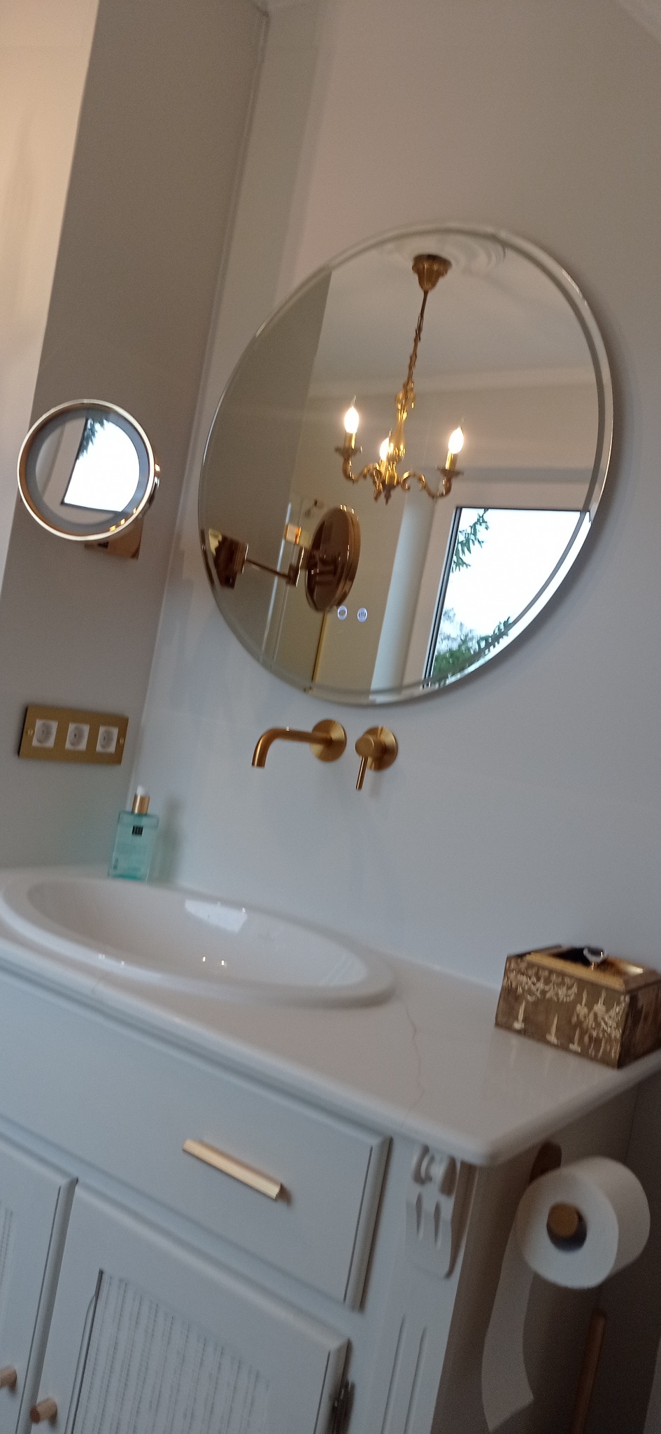 Elegancka łazienka z okrągłym lustrem, złotą armaturą i szafką pod umywalką w stylu retro. W lustrze odbija się żyrandol, dodając wnętrzu luksusowego charakteru.