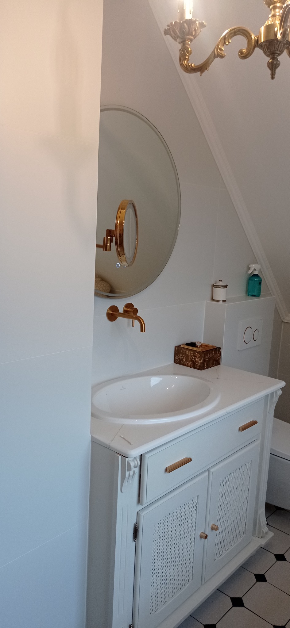 Biała szafka umywalkowa z marmurowym blatem i złotą armaturą w łazience na poddaszu. Okrągłe lustro z podświetleniem i dekoracyjny żyrandol.