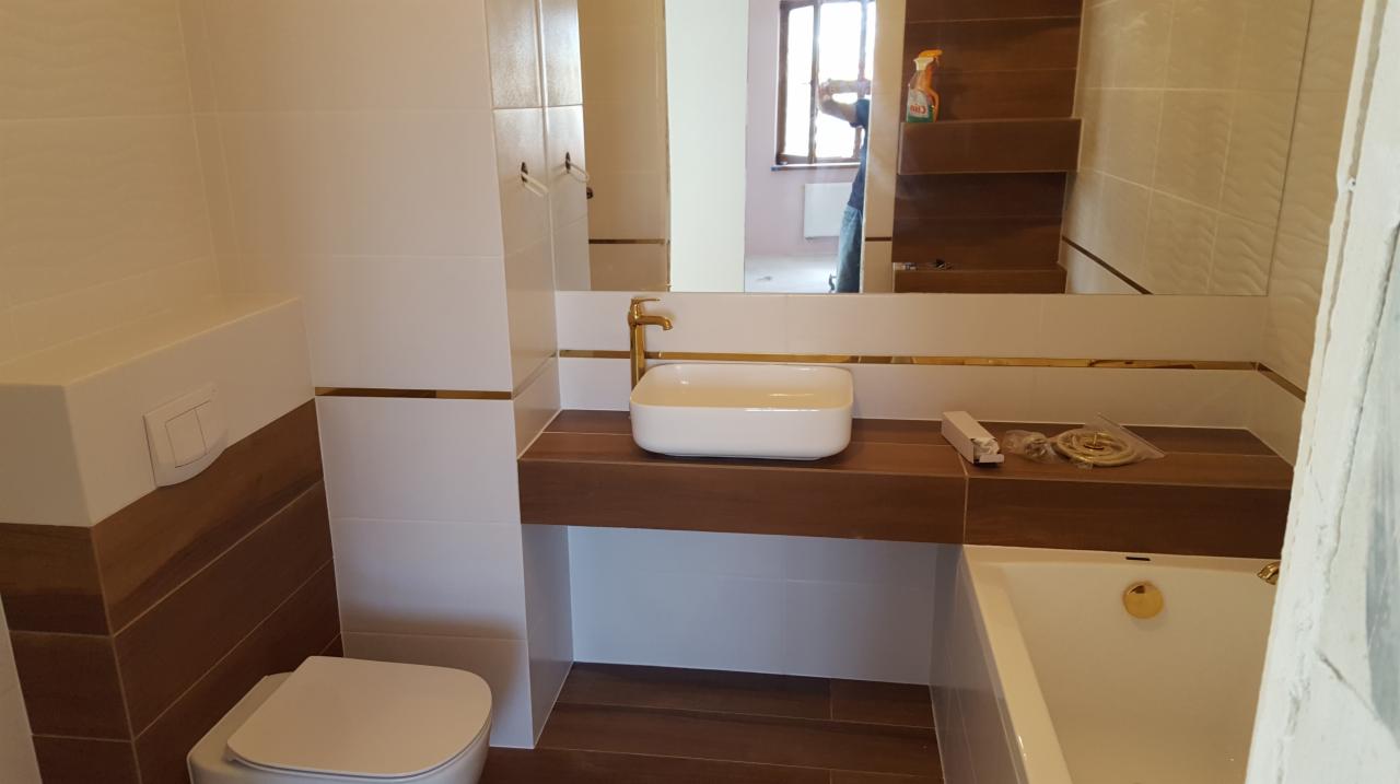 Nowoczesna łazienka z białymi i brązowymi płytkami, złotymi akcentami, białą umywalką nablatową i widoczną w lustrze osobą wykonującą prace wykończeniowe.