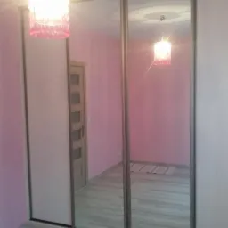 Wnętrze pokoju z trzydrzwiową szafą przesuwną z lustrzanymi frontami, odbijającymi drzwi i dywanik, oraz wiszącą lampą z różowymi elementami.