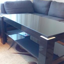 Meble na wymiar Art Arco Arkadiusz Kulesza - Stolik kawowy z ciemnego drewna z szklanym blatem i półką, ustawiony na beżowym dywanie w brązowe koła, w tle szara sofa i biała zasłona.