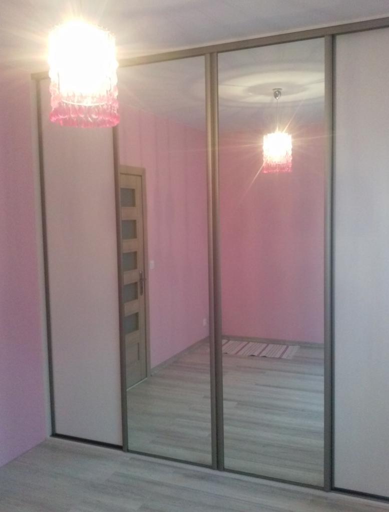 Wnętrze pokoju z trzydrzwiową szafą przesuwną z lustrzanymi frontami, odbijającymi drzwi i dywanik, oraz wiszącą lampą z różowymi elementami.