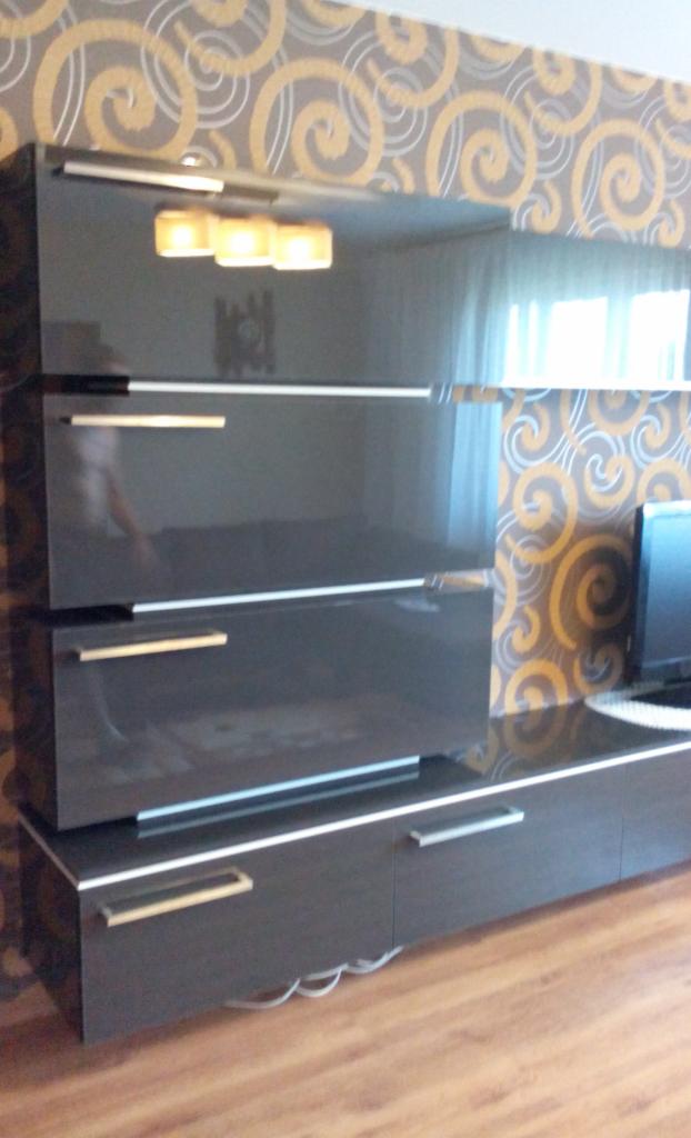 Ciemnoszara, błyszcząca komoda z szufladami i złotymi uchwytami na tle ściany z wzorzystą tapetą, widoczny fragment podłogi z paneli.