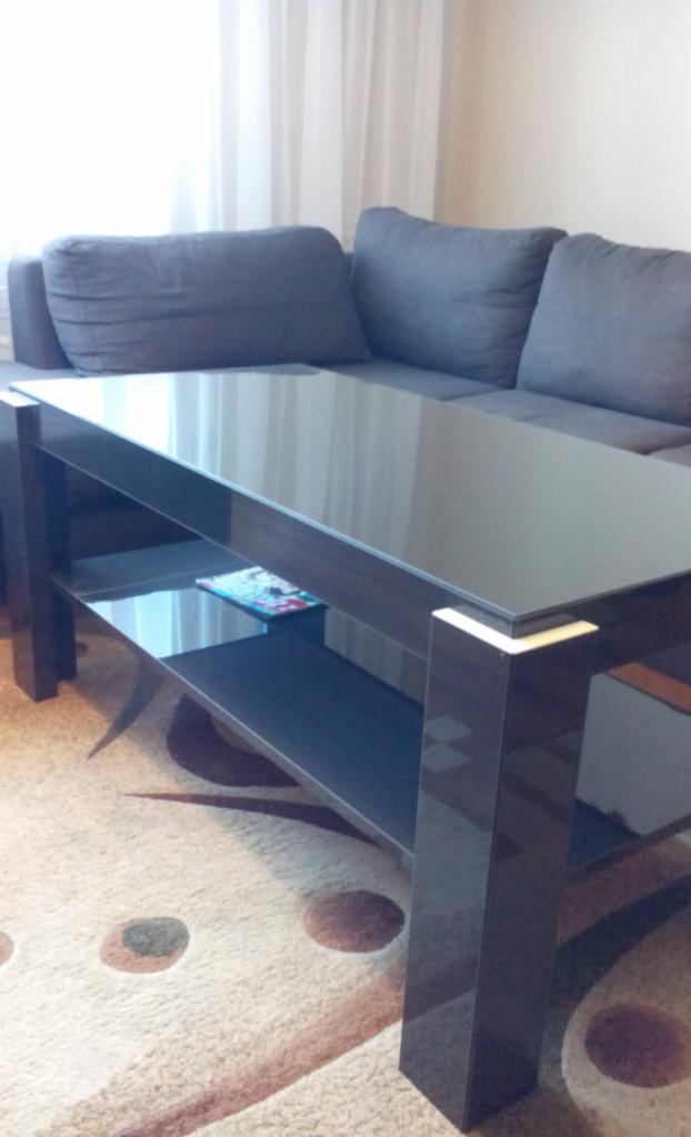 Stolik kawowy z ciemnego drewna z szklanym blatem i półką, ustawiony na beżowym dywanie w brązowe koła, w tle szara sofa i biała zasłona.