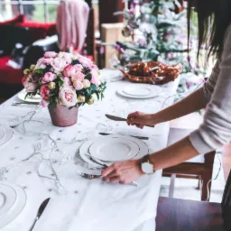 Ustawianie stołu: kobieta układa sztućce obok białych talerzy z złotym obramowaniem na białym obrusie w gwiazdki, dekoracje świetlne, bukiet róż w miedzianym wazonie, choinka w tle.
