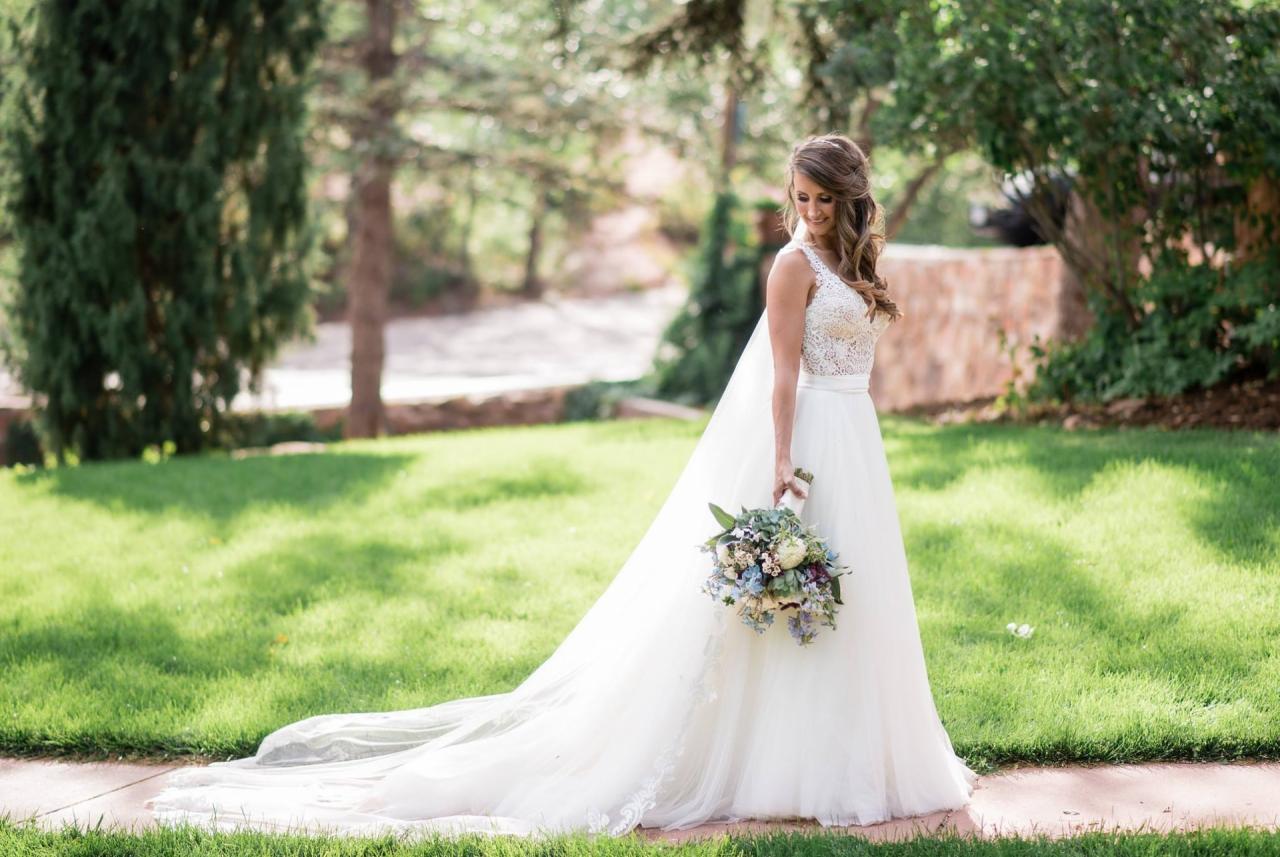 Uśmiechnięta panna młoda w koronkowej sukni z długim trenem i bukietem, pozuje na tle zielonego trawnika i drzew w słoneczny dzień.