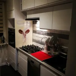 Biała kuchnia z błyszczącymi frontami, szare płytki na ścianie, płyta gazowa i zmywarka; na panelu nad płytą grafika z motywem kawy.