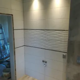 Ściana łazienki wyłożona białymi płytkami z poziomym pasem dekoracyjnych płytek o falistym wzorze, widoczne chromowane zawory i gniazdko elektryczne, w tle drabina i wiadro budowlane.