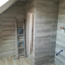 Ukończona łazienka na poddaszu z szaro-beżowymi płytkami imitującymi drewno na ścianach i podłodze, widoczna drabina malarska i niebieska taśma malarska wzdłuż krawędzi sufitu.
