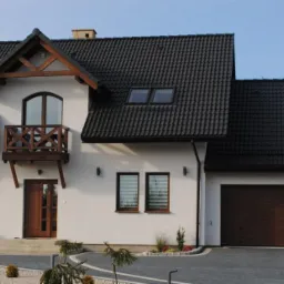 Nowoczesny dom jednorodzinny z białymi ścianami, ciemną dachówką i brązowymi ramami okiennymi oraz drewnianym balkonem, otoczony zadbanym ogrodem z kostki brukowej.