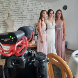 Kamera z mikrofonem nagrywa trzy kobiety ubrane w eleganckie suknie, prawdopodobnie podczas przygotowań do ślubu, na tle białej ceglanej ściany i manekina krawieckiego.