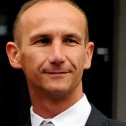 Portret mężczyzny w garniturze i krawacie w odcieniach szarości, z uśmiechem na twarzy i krótkimi włosami, na rozmytym tle.