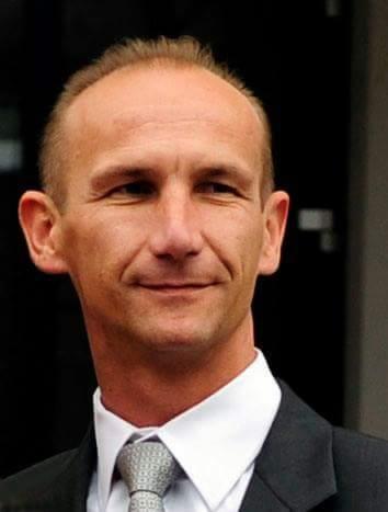 Portret mężczyzny w garniturze i krawacie w odcieniach szarości, z uśmiechem na twarzy i krótkimi włosami, na rozmytym tle.