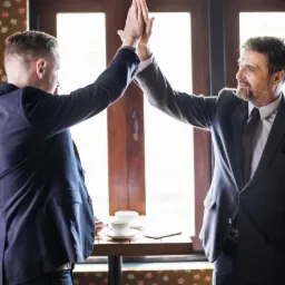 Dwóch mężczyzn w garniturach daje sobie 'piątkę' nad stolikiem z filiżankami kawy i tabletem, w tle okno z drewnianymi ramami.