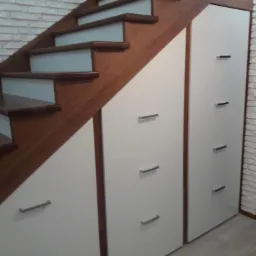 Drewniane schody zintegrowane z białymi szafkami z podłużnymi, metalowymi uchwytami, umieszczone przy ścianie z dekoracyjnej białej cegły.