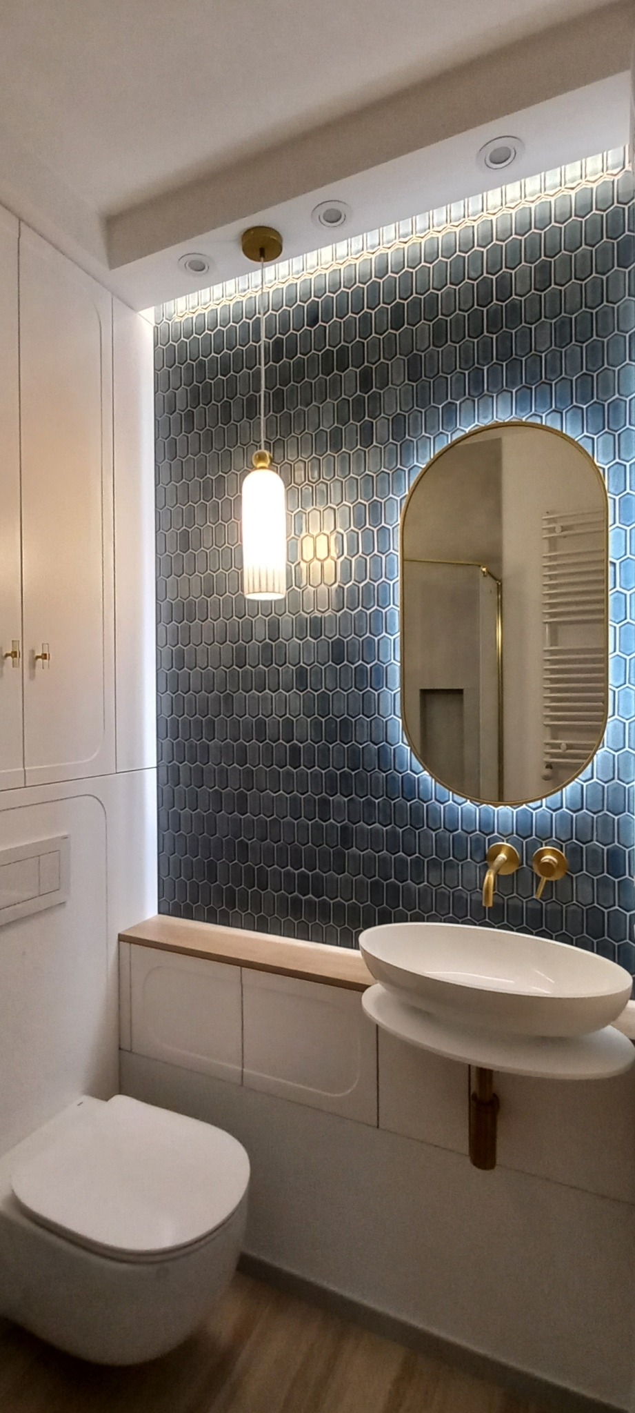Nowoczesna łazienka z umywalką nablatową, podświetlanym lustrem owalnym z ramą w kolorze złota, armaturą w kolorze złota, płytkami heksagonalnymi w odcieniach niebieskiego i białymi szafkami.