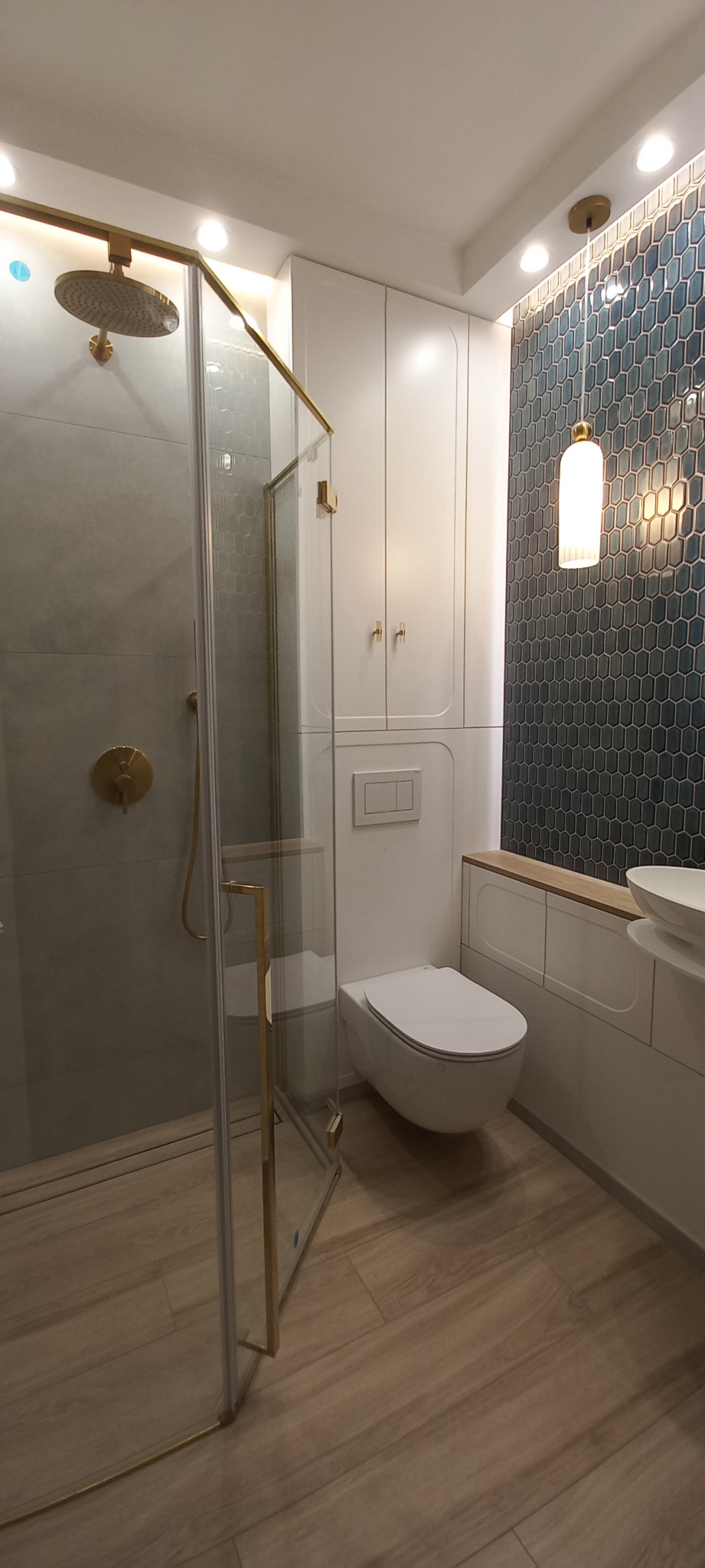 Nowoczesna łazienka z kabiną prysznicową typu walk-in z złotymi elementami, wiszącą toaletą, szafkami w zabudowie i ścianą pokrytą niebieską mozaiką w kształcie plastra miodu.