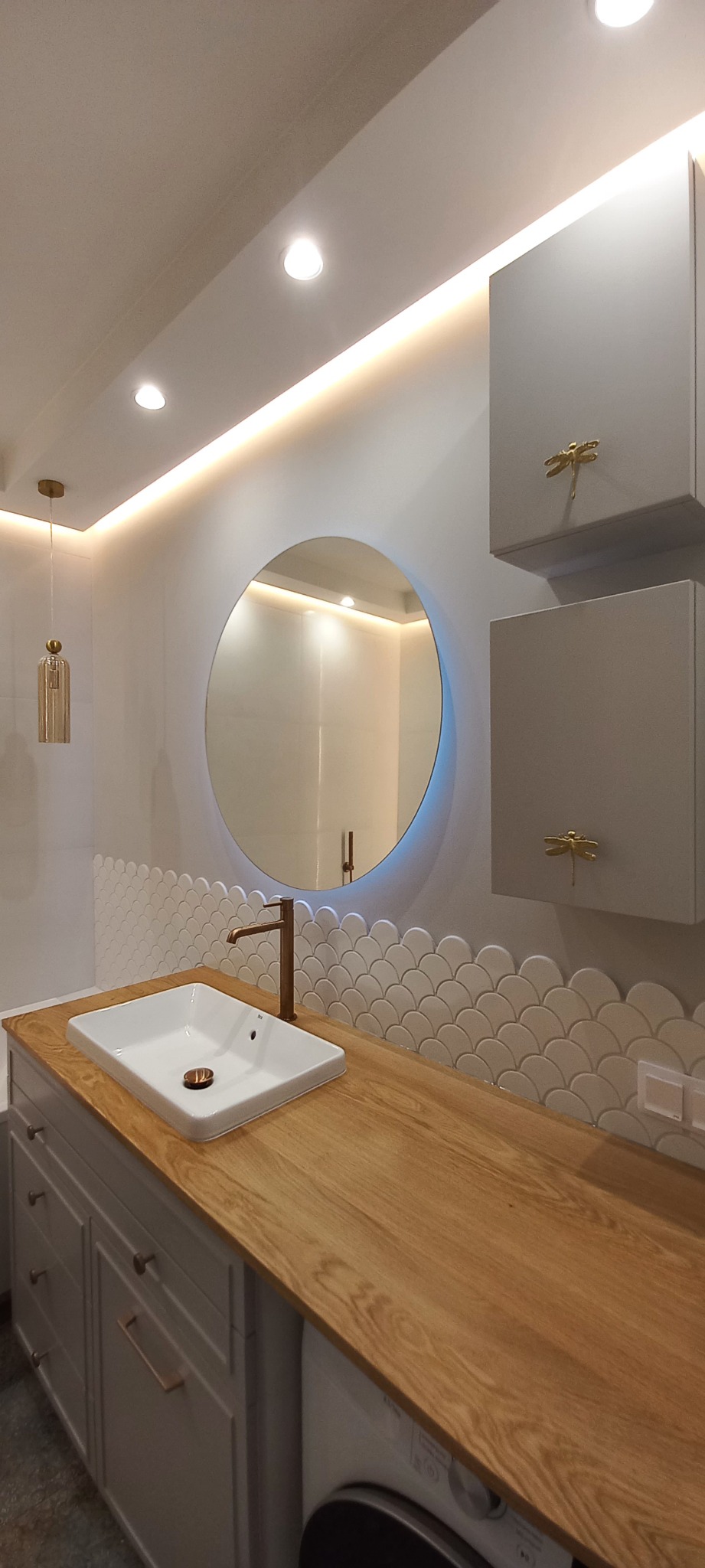 Nowoczesna łazienka z okrągłym lustrem podświetlanym LED, umywalką nablatową z brązową armaturą, drewnianym blatem i szafkami z dekoracyjnymi uchwytami w kształcie ważek.