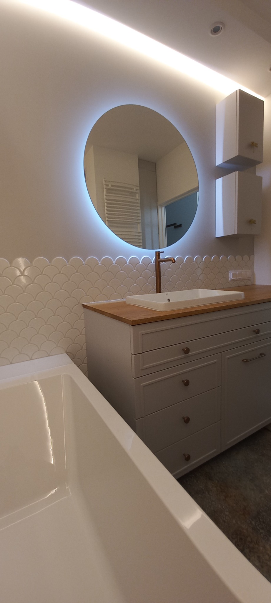 Nowoczesna łazienka z okrągłym lustrem podświetlanym LED, białą umywalką na drewnianym blacie, szafkami wiszącymi i wanną z białymi płytkami w kształcie rybiej łuski na ścianie.