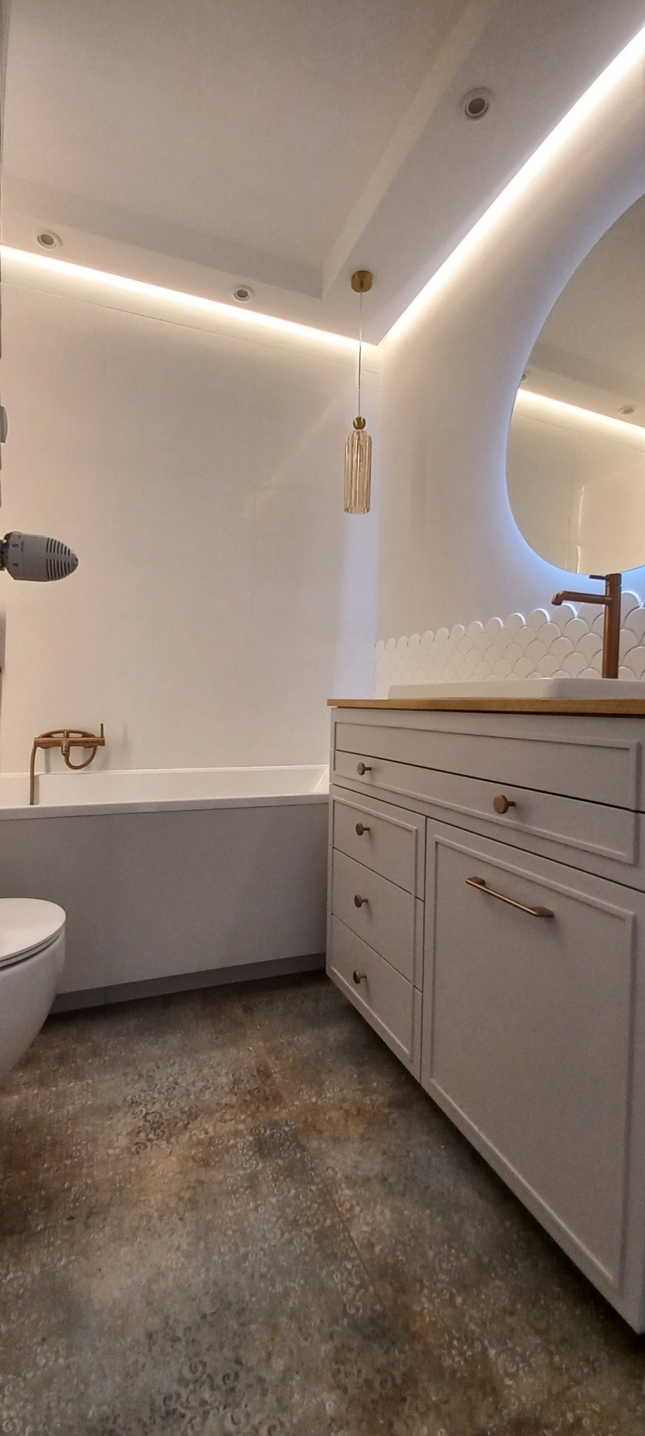 Nowoczesna łazienka z wanną, umywalką na szafce z drewnianym blatem, okrągłym lustrem z podświetleniem LED i wiszącą lampą w stylu glamour.