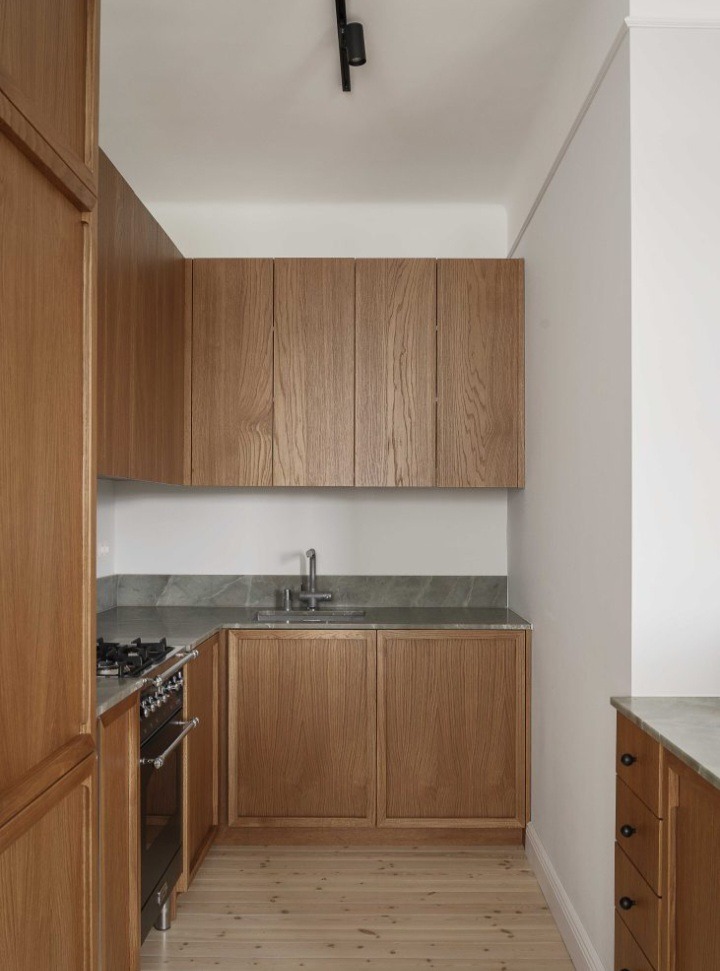 Wąska kuchnia z drewnianymi frontami szafek, szarym blatem i jasną drewnianą podłogą, oświetlona reflektorem sufitowym.