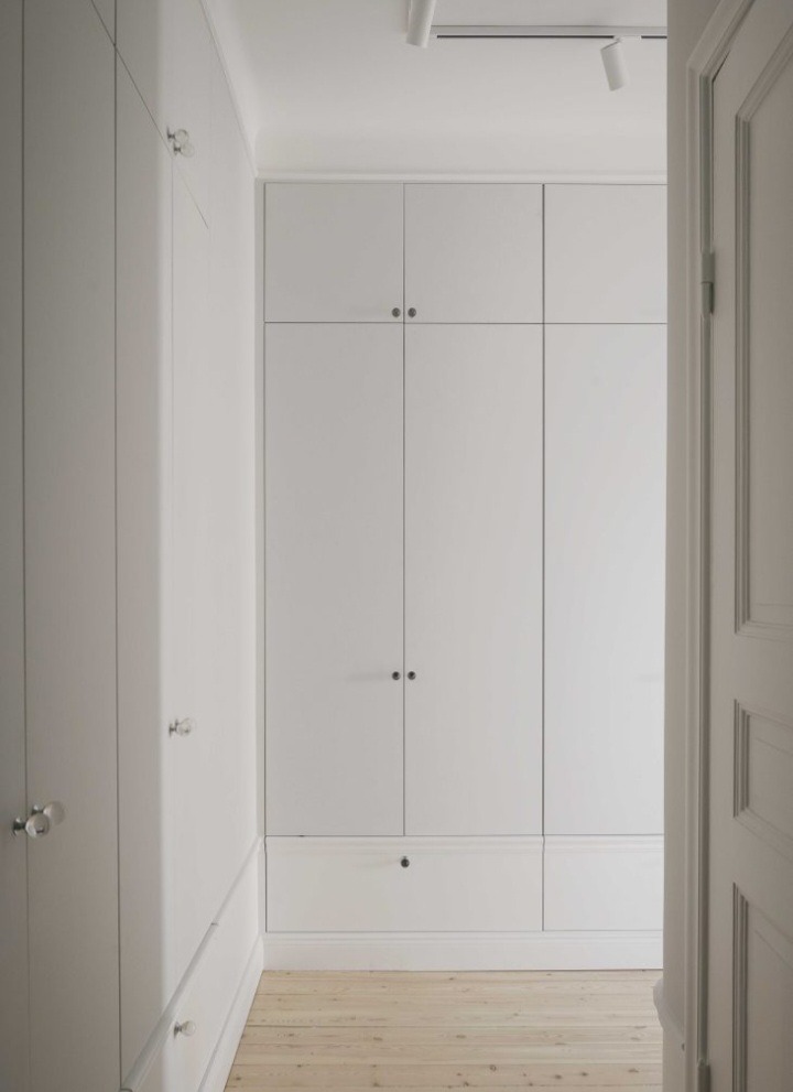 Biała, minimalistyczna zabudowa szaf w przedpokoju z drewnianą podłogą, widok zza uchylonych drzwi.