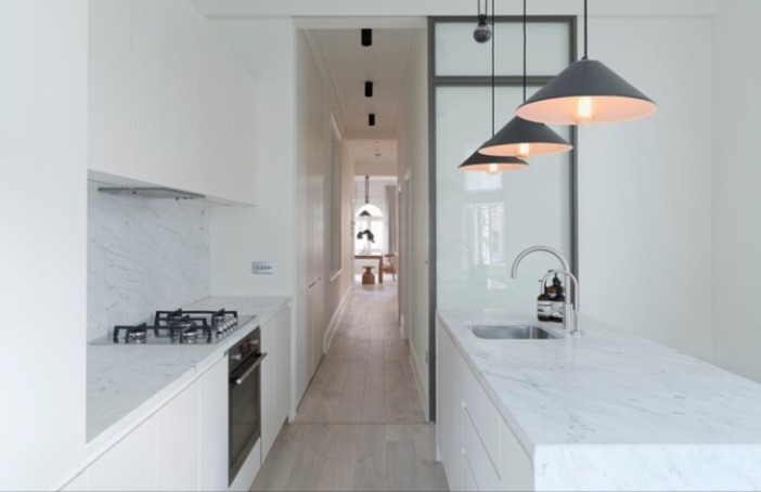 Wąska, minimalistyczna kuchnia w bieli z marmurowym blatem, czarnymi lampami stożkowymi i widokiem na dalszą część mieszkania.