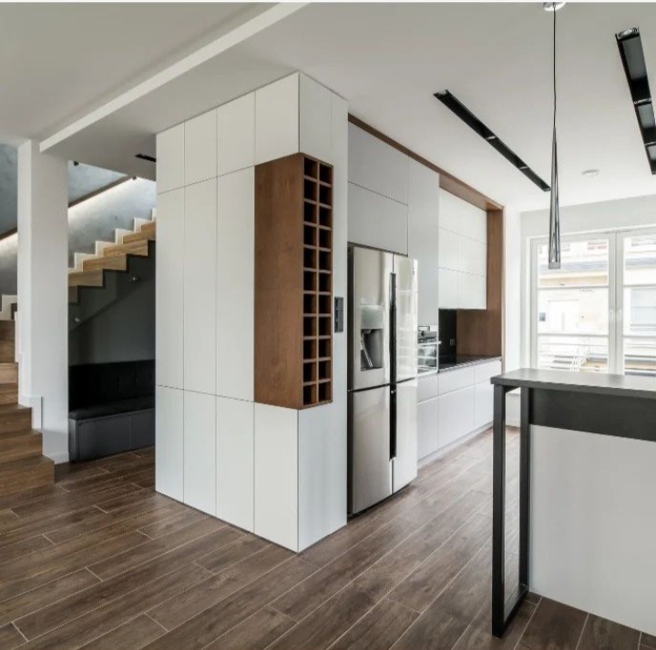 Nowoczesna kuchnia z białymi szafkami, drewnianym regałem na wino i stalową lodówką, widok na drewniane schody i minimalistyczne oświetlenie.