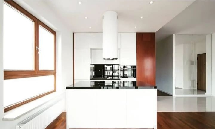 Nowoczesna kuchnia z białymi szafkami, czarnym blatem i cylindrycznym okapem, drewniana podłoga i okno z brązową ramą.