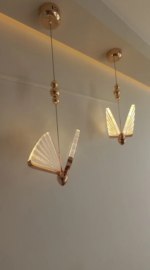 Dwa wiszące, złote, dekoracyjne żyrandole w kształcie motyli z przezroczystymi, żłobionymi skrzydłami, oświetlające sufit.