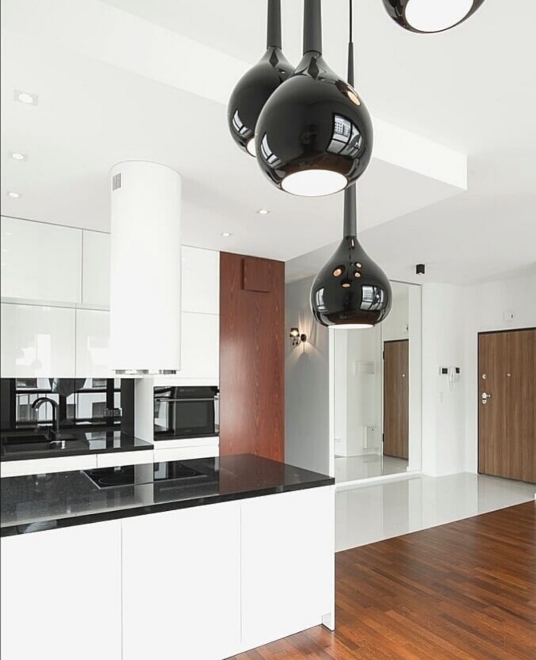Nowoczesna kuchnia z białą zabudową, czarnym blatem i błyszczącymi, kulistymi lampami wiszącymi; parkiet z ciemnego drewna.
