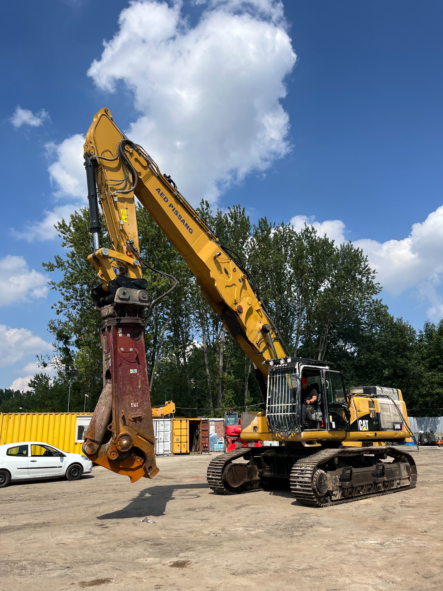 Żółta koparka gąsienicowa CAT 349D z hydraulicznym nożycami do betonu AED PISSANG, na placu budowy, w tle kontenery i drzewa, błękitne niebo z chmurami.