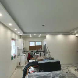 Wnętrze w trakcie odświeżania: świeżo pomalowane ściany na biało, widoczne przygotowania do dalszych prac wykończeniowych, zabezpieczone meble, oświetlenie sufitowe.